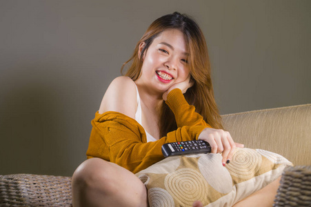 年轻快乐迷人的亚洲女性在家看电视沙发沙发遥控微笑放松享受喜剧电影或电视节目的家庭娱乐理念