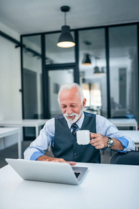 快乐的高级商人在办公室里使用笔记本电脑时喝咖啡。