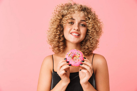 20岁的漂亮卷发女士穿着连衣裙，一边吃甜甜圈，一边站在粉红色的背景上