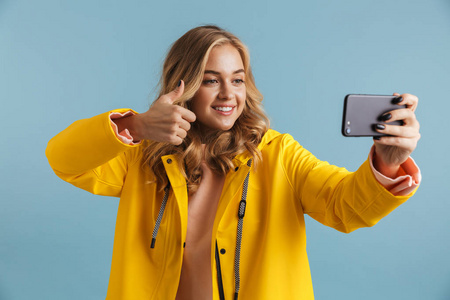 欧洲20岁的女人穿着黄色雨衣,拿着手机,在蓝色背景下拍摄自拍照片照片