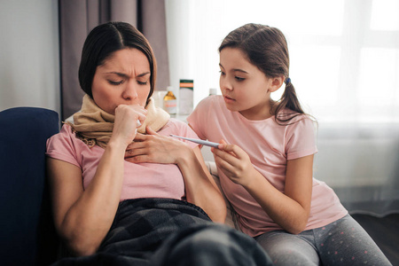 年轻女子咳嗽。她用拳头捂住嘴。褐发女郎和她的女儿坐在沙发上在房间里。她看着孩子们手里的温度计。女孩拥抱母亲