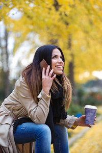 一个美丽的年轻女人在手机上说话，一边喝咖啡，一边坐在街上的长凳上。