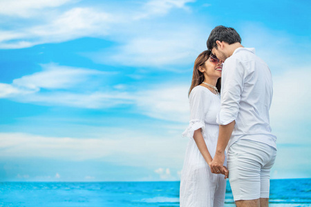 年轻一对相爱的年轻夫妇互相看着对方，手牵着手在蓝天上的海边。快乐的微笑年轻的婚礼，白色的连衣裙，亲吻浪漫的时刻，从头到脚或面对面