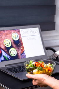 女性手中的蔬菜沙拉碗靠近桌面上的笔记本电脑。在屏幕上计划从星期一开始减肥