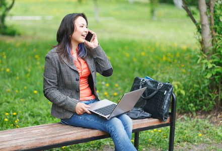 年轻的自由职业者女孩坐在公园的长凳上，用笔记本电脑和电话交谈