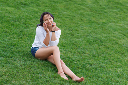 快乐美丽的年轻女子坐在草地上，用智能手机