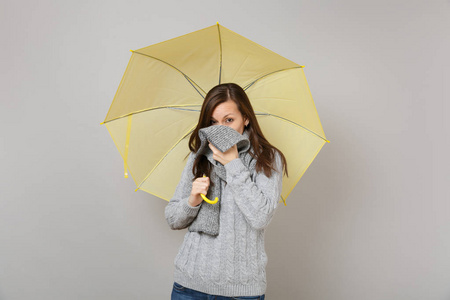 年轻女子穿着灰色毛衣，遮住嘴脸，围巾拿着黄色的伞，孤立在灰色的墙壁背景上。 健康时尚生活方式人们情感冷季观念。 模拟复制空间