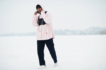 时尚的城市风格非裔美国人穿着粉红色连帽衫在冬天的冰冻湖上摆姿势。