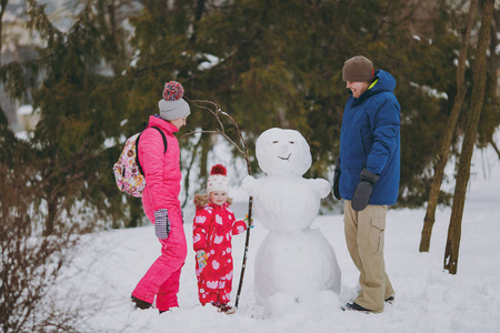 幸福的年轻家庭，男人，女人，穿着温暖的衣服，站在雪地公园或森林户外的雪人附近。 假日的冬季乐趣休闲。 家庭生活方式观念