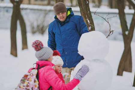年轻的家庭男人女人小女孩穿着冬天温暖的衣服在雪地公园或户外森林里堆雪人。 假日的冬季乐趣休闲。 家庭生活方式观念