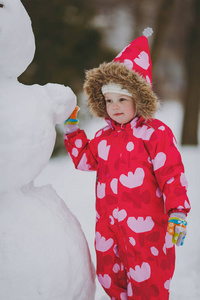 漂亮的小女孩穿着冬天温暖的衣服，戴着兜帽，站在公园或森林户外的雪人附近。 假日的冬季乐趣休闲。 恋爱关系家庭童年生活方式