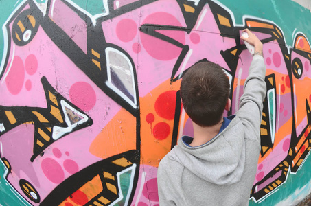 一个穿灰色连帽衫的年轻人在雨天在墙上画粉红色和绿色的涂鸦。 菲什耶枪