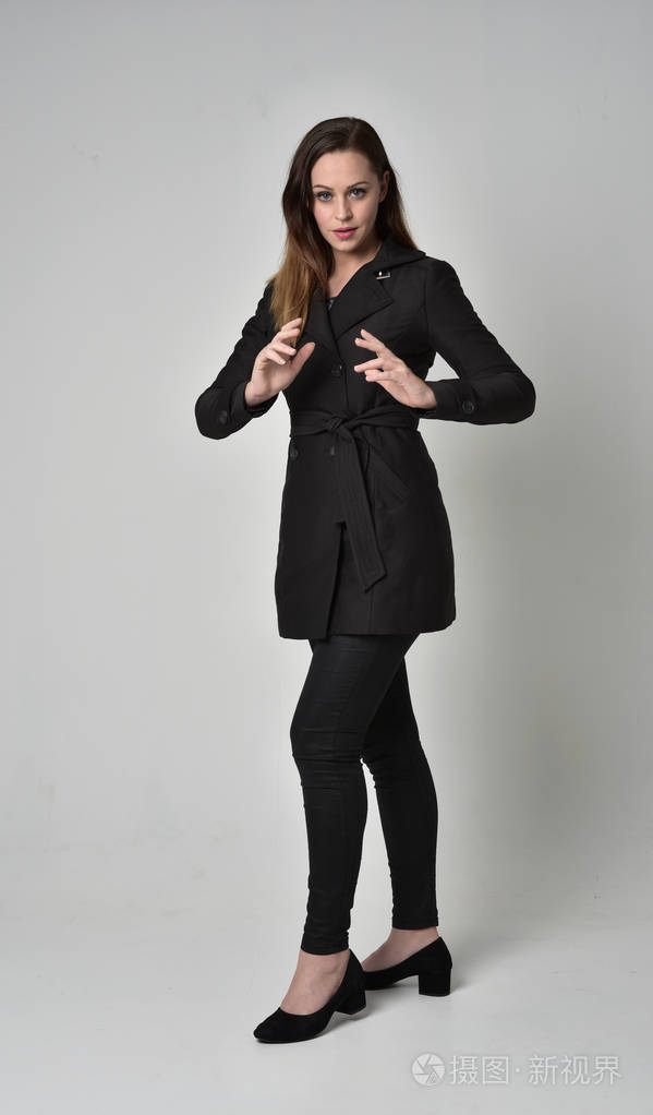 一个黑发女孩穿着黑色长外套站在灰色工作室背景上的姿势的全长肖像.
