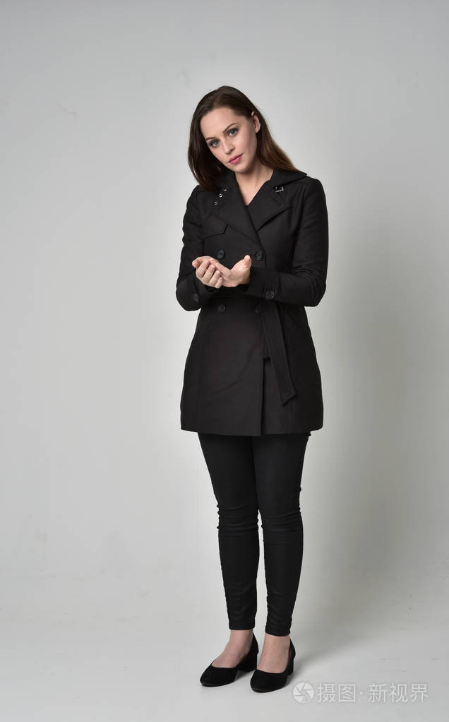 一个黑发女孩穿着黑色长外套站在灰色工作室背景上的姿势的全长肖像