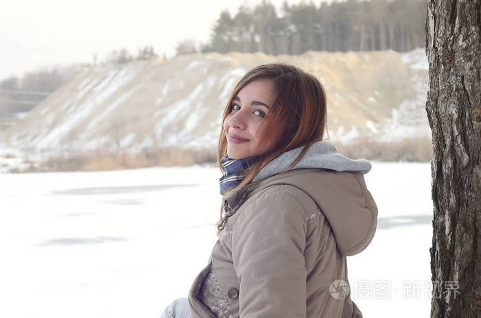 一个穿着棕色外套的年轻白种人女孩正坐在悬崖附近，在冬天，天空和冰冻的湖泊之间有一条地平线。