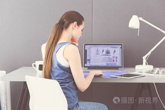 年轻的女人坐在办公桌前与文书, 计划和笔记本电脑