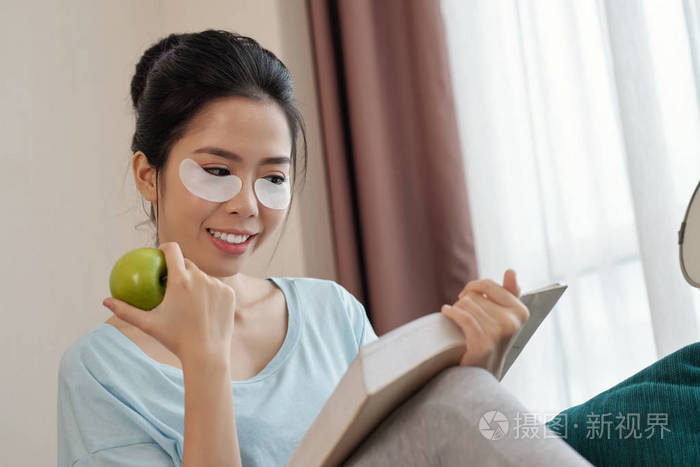 可爱的亚洲女士，成熟的苹果和眼垫下，微笑着阅读有趣的书，同时放松客厅