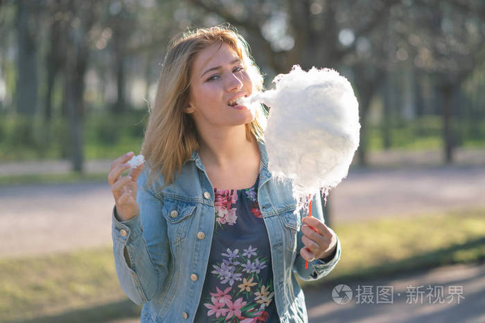 迷人的白种人女孩穿着牛仔夹克在春天公园散步,在早晨的阳光下吃着粉