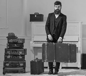 男子气概优雅, 在严格的脸上站在一堆老式手提箱, 持有手提箱。男人, 带着胡子和胡须的旅行者带着行李, 奢华的白色内饰背景。行李