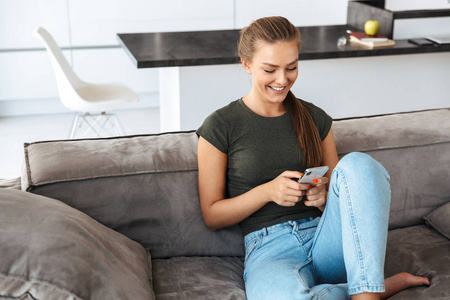 开朗的年轻女人在家坐在沙发上用手机