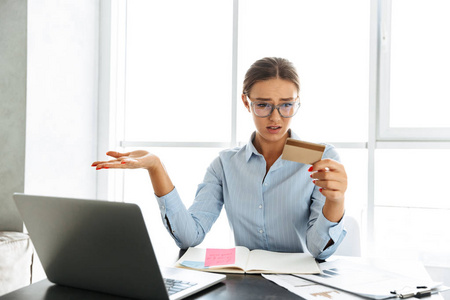 困惑的女商人坐在厨房里拿着笔记本电脑看塑料信用卡