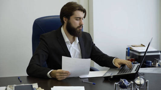 英俊的商人在办公室里用笔记本电脑工作。留着胡子在办公室的电脑前工作的年轻商人