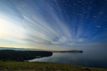 俄罗斯贝加尔湖奥尔洪岛云和星星在一个月亮的夜晚在小海湾上空。 星星的短暂褪色轨迹在云层中回响