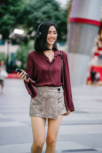 美丽的韩国女人穿着优雅的衬衫和短裤跳舞，她走在城市的一条街上，戴着耳机