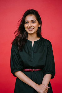 一个美丽的年轻和有吸引力的印度亚洲妇女在红色工作室背景下的工作室肖像。 她优雅地穿着一件简单的绿色连衣裙和红色腰带，微笑着。