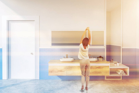 女性在现代浴室的内部，白色和蓝色砖墙，混凝土地板和双槽站在木制台面上，水平和垂直镜子。 调色图像