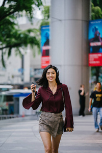 美丽的韩国女人穿着优雅的衬衫和短裤跳舞，她走在城市的一条街上，戴着耳机