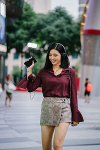 美丽的韩国女人穿着优雅的衬衫和短裤跳舞，她走在城市的一条街上，戴着耳机