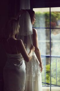 漂亮的女孩或伴娘帮助美丽的新娘的女人穿着白色婚纱，把蕾丝面纱放在室内的窗户背景上