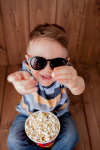 小可爱的孩子男孩2，3岁的3DIMA X影院眼镜，拿着桶爆米花，在木制背景下吃快餐。 儿童童年生活方式的概念。 复制空间。