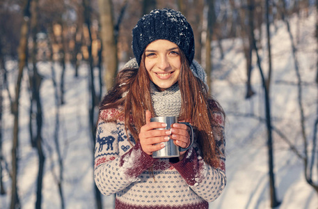 微笑的漂亮女人，喝着一杯热饮料，站在冬季森林附近，在令人惊叹的冬季景观中度假。 流浪的风格