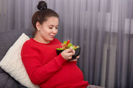 侧镜头高兴准妈妈吃新鲜素食沙拉拿叉子有发结穿着随意坐在舒适的沙发靠近窗户享受健康的饮食和生活方式。