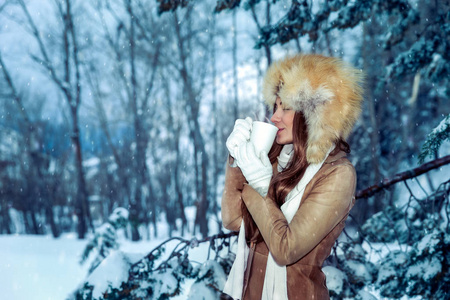 平静满足满足的女人高兴地在白雪覆盖的树木中喝热咖啡，享受在公园散步的寒假