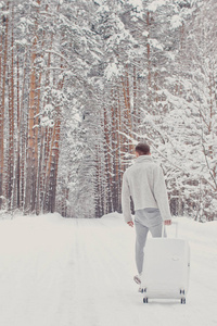 年轻时尚人士的肖像，穿着白色衣服，走在雪地上，拿着手提箱，走在松林的小径上。 寒假旅行的概念。 冬天在户外。 滑雪场，大型滑雪场