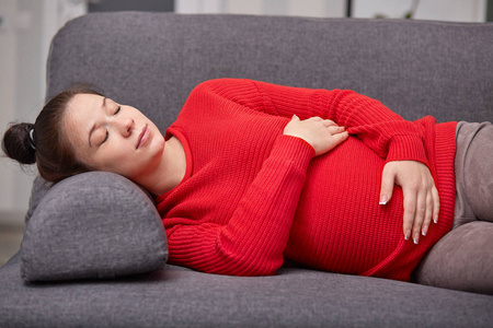 睡累孕妇午睡照片,双手放在肚子上,穿红色温暖的毛衣,在家里的沙发上