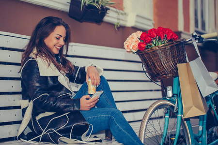 快乐的年轻女子坐在城市街道的长凳上，一边用手机看短信，一边喝咖啡