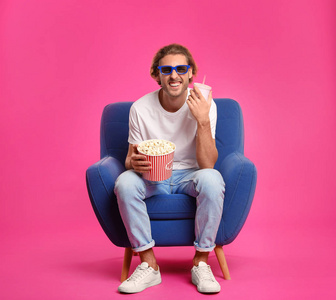 3D眼镜爆米花和饮料的男人坐在扶手椅上观看彩色背景电影