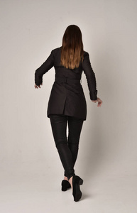 一个穿着黑色长外套的深色女孩的全长肖像，站在灰色工作室背景上的镜头后面。