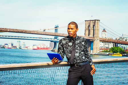非裔美国商人在纽约旅行。 穿着黑色的花图案衬衫，系着一个年轻人站在港口读蓝色平板电脑。 曼哈顿布鲁克林大桥的背景