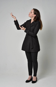 一个黑发女孩穿着黑色长外套，站在侧面轮廓灰色工作室背景的完整长度肖像。