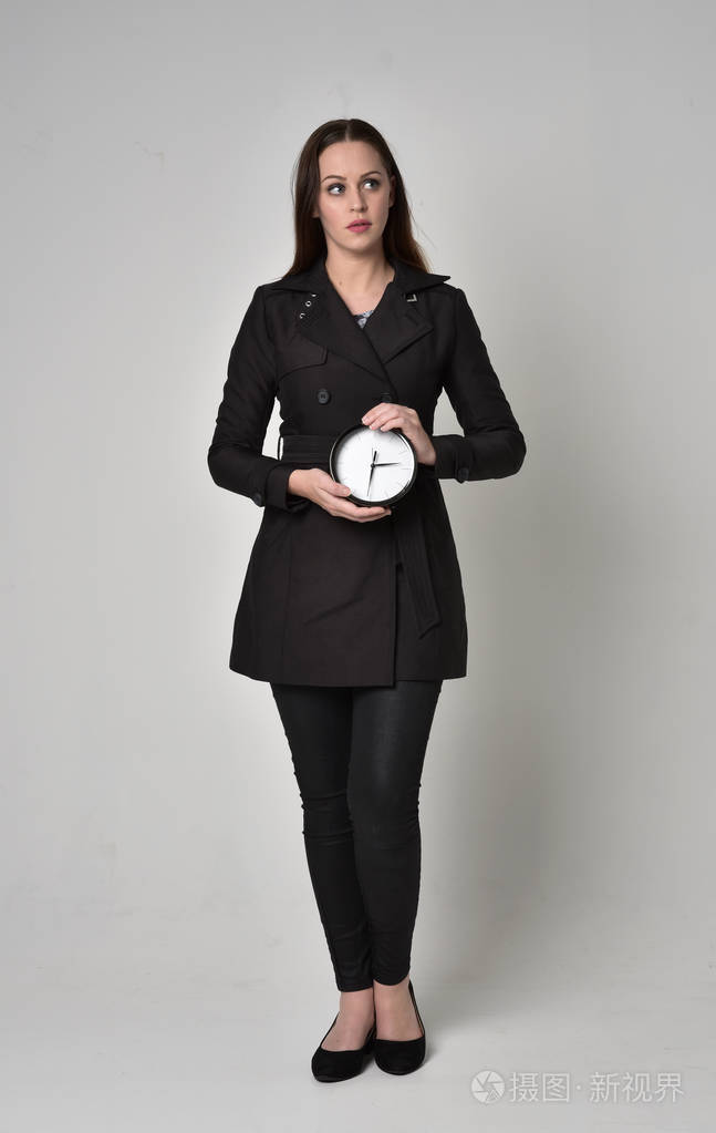 一个黑发女孩穿着黑色长外套站在灰色工作室背景上的姿势的全长肖像