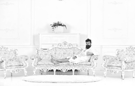 男子昏昏欲睡的浴衣喝咖啡在豪华酒店在早上, 白色的背景。奢华生活理念。胡子和胡子的人享受早晨, 而坐在老式的豪华沙发上