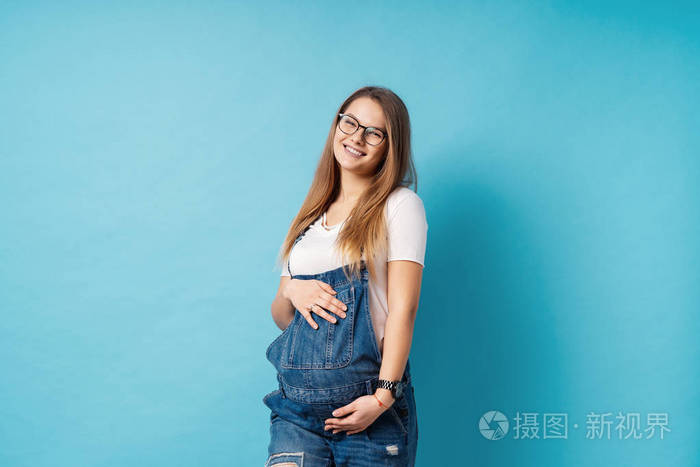 一个可爱的年轻女人戴着眼镜和牛仔裤,摆着摸肚子,与她未出生的孩子