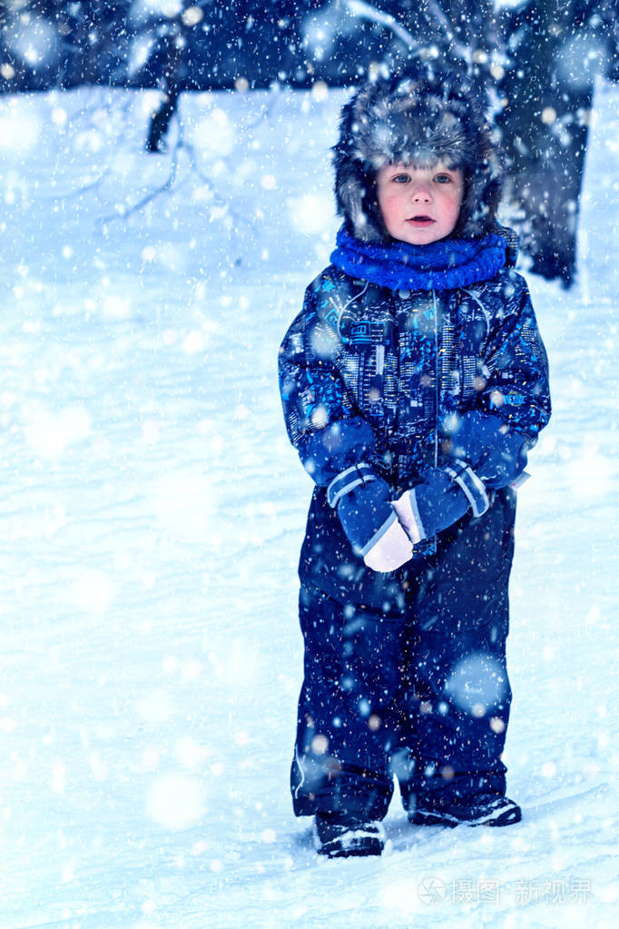 下雪时穿着工作服和毛皮帽子衣服在户外玩耍的有趣的小男孩.