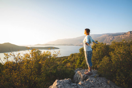 快乐的旅行者，张开双臂的女人站在岩石上，在日落时欣赏地中海的绿松石水域沿着土耳其的莱西亚路冒险旅行。后视，背光..