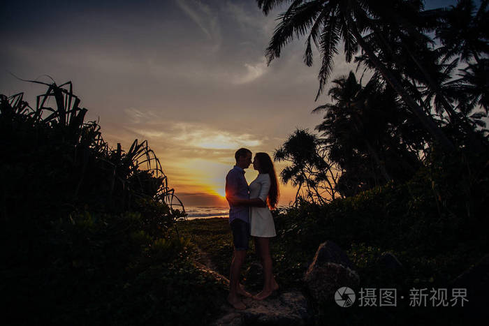 两个年轻的恋人站在海滩上在日落的棕榈树上互相看着